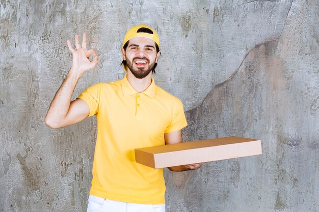 Courier in yellow uniform holding a takeaway pizza box and showing positive hand sign