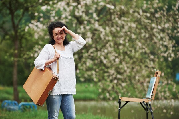 Covering eyes by hand from sunlight. Mature painter with case of instruments have a walk in the beautiful spring park