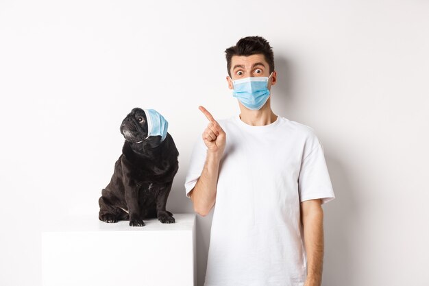 코비드-19, 동물 및 검역 개념. 개 주인과 의료용 마스크를 쓴 귀여운 검은 퍼그, 가리키는 남자, 왼쪽 위 모서리, 흰색 배경을 쳐다보는 강아지.