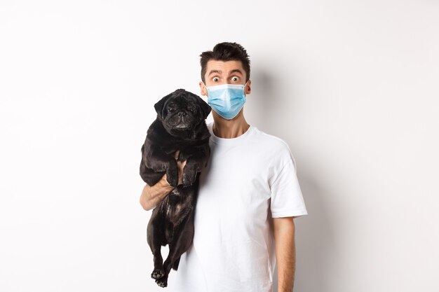 Covid-19, животные и концепция карантина. Забавный молодой человек в медицинской маске, держащий милый черный мопс, стоящий на белом фоне