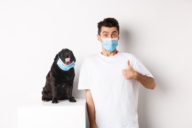Covid-19、動物と検疫の概念。面白い若い男と医療マスクの小さな犬の画像、承認で親指を示す所有者、何かを賞賛、白い背景