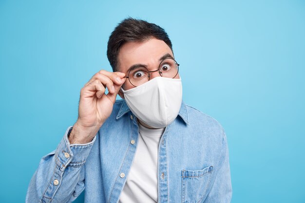 Covid 19 concept. Serious attentive adult man looks through spectacles wears protective face mask asks to follow social distancing rules dressed in denim shirt 