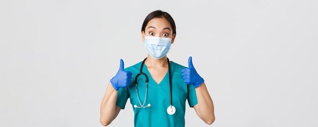 Covid-19, coronavirus disease, healthcare workers concept. Amused smiling asian doctor, physician in medical mask and rubber gloves, showing thumbs-up, approve, praise nice work.