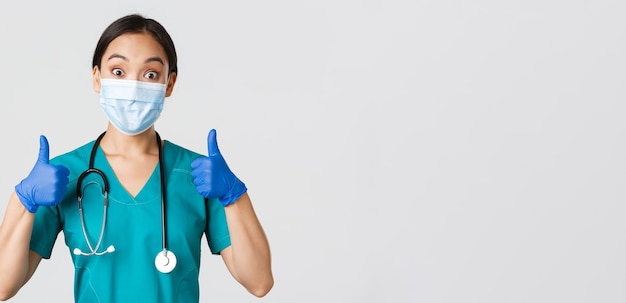 Covid-19, coronavirus disease, healthcare workers concept. Amused smiling asian doctor, physician in medical mask and rubber gloves, showing thumbs-up, approve, praise nice work