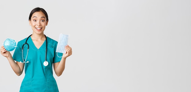 Covid-19, coronavirus disease, healthcare workers concept. Amused smiling asian female nurse, inters in scrubs showing respirator and medical mask, receive personal protective equipment.