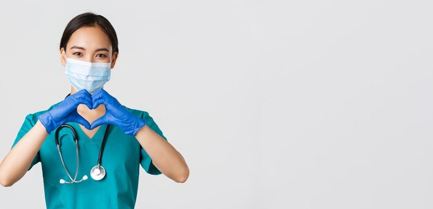 Ковид-19, коронавирусная болезнь, концепция медицинских работников. Крупный план очаровательной улыбающейся азиатской женщины-врача, врача в медицинской маске и резиновых перчатках, ухаживающей за пациентами, показывающей сердечный жест.