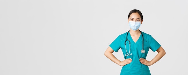 Covid-19, coronavirus disease, healthcare workers concept. Confident professional physician, doctor in medical mask and scrubs working her shift clinic, looking determined, white background