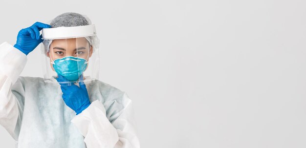 Covid-19, coronavirus disease, healthcare workers concept. Confident serious-looking female asian doctor, put on face shield and respirator, personal protective equipment, white background