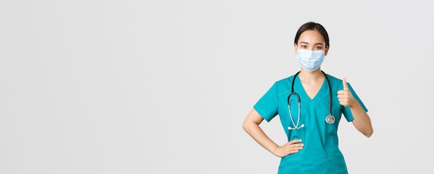 Free photo covid-19, coronavirus disease, healthcare workers concept. confident smiling asian doctor, female nurse or physician in medical mask and scrubs, show thumbs-up assure quality, guanratee service