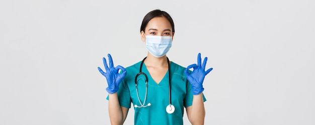 Free photo covid-19, coronavirus disease, healthcare workers concept. confident smiling asian female doctor, nurse in medical mask and gloves, show okay gesture in approval, white background.