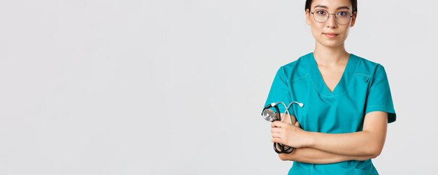 Covid-19, coronavirus disease, healthcare workers concept. Confident smiling professional asian doctor in glasses, cross arms chest, wearing scrubs and holding stethoscope, white background.