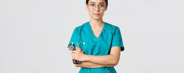 Foto gratuita covid-19, malattia da coronavirus, concetto di operatori sanitari. medico asiatico professionista sorridente sicuro con gli occhiali, petto a braccia incrociate, scrub da indossare e stetoscopio in mano, sfondo bianco