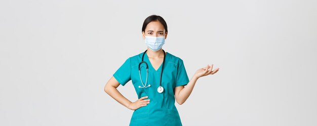 Covid-19, coronavirus disease, healthcare workers concept. Confused and upset asian female doctor in medical mask and scrubs, raise hand up puzzled, cant understand what problem, white background.
