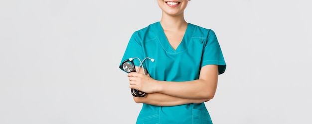Ковид-19, коронавирусная болезнь, концепция медицинских работников. Обрезанный снимок тела азиатской женщины-врача, улыбающейся, скрещенной на груди уверенно и держащей стетоскоп, стоящей на белом фоне