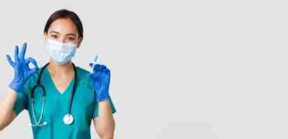 Free photo covid-19, coronavirus disease, healthcare workers concept. cute asian female doctor, physician in medical mask and rubber gloves show okay gesture and hold syringe with vaccine, white background.