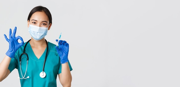 Covid-19, coronavirus disease, healthcare workers concept. Cute asian female doctor, physician in medical mask and rubber gloves show okay gesture and hold syringe with vaccine, white background.