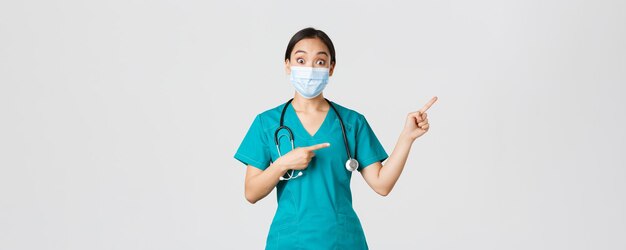 Covid-19, coronavirus disease, healthcare workers concept. Enthusiastic happy asian female doctor, physician or nurse in medical mask and scrubs, pointing upper right corner, show awesome promo