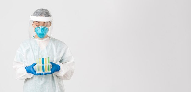 Covid-19, coronavirus disease, healthcare workers concept. Excited asian female doctor, tech lab in personal protective equipment holding test-tubes with vaccine or analyses, white background.