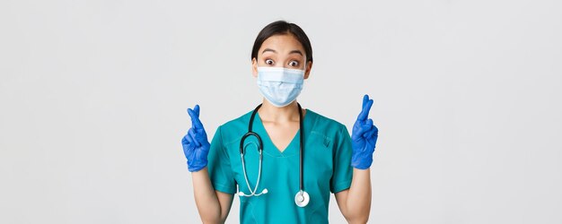 Covid-19、コロナウイルス病、医療従事者の概念。興奮して希望に満ちたアジアの女性医師、医療マスクとゴム手袋の看護師は、幸運、makignの願い、白い背景の指を交差させます