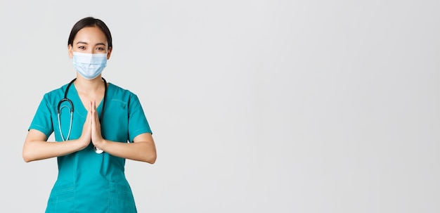 Covid-19, коронавирусная болезнь, концепция медицинских работников. Обнадеживающая улыбающаяся азиатская женщина-врач, врач в медицинской маске и скрабах держатся за руки в молитвенном жесте, благодаря, на белом фоне.