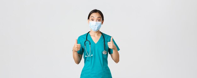 Ковид-19, коронавирусная болезнь, концепция медицинских работников. Впечатленная счастливая азиатская женщина-врач, врач в медицинской маске и скрабах показывает палец вверх, соглашается, хвалит выбор, хвалит хорошую работу.