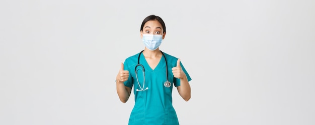 Covid-19, coronavirus disease, healthcare workers concept. Impressed happy asian female doctor, physician in medical mask and scrubs show thumbs-up, agree, compliment choice, praise nice work