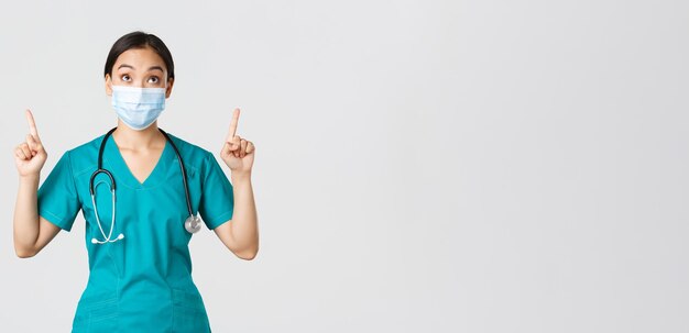 Covid-19, coronavirus disease, healthcare workers concept. Intrigued and excited asian woman doctor, nurse in medical mask and gloves, looking and pointing fingers up, white background