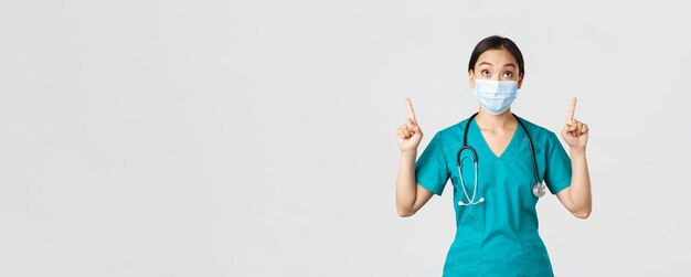 Covid-19、コロナウイルス病、医療従事者の概念。興味をそそられ、興奮しているアジアの女性医師、医療マスクと手袋の看護師、指を上に向けて見ている、白い背景