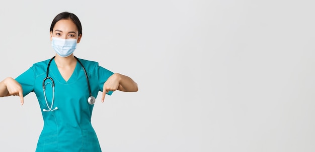 Free photo covid-19, coronavirus disease, healthcare workers concept. pleasant smiling asian female nurse, doctor in medical mask and scrubs, pointing fingers down, provide information, invite for checkup