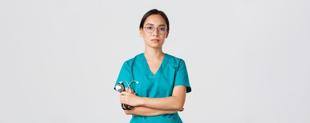 Covid-19, coronavirus disease, healthcare workers concept. Professional confident, serious-looking female doctor, physician in glasses and scrubs, holding stethoscope, standing white background.