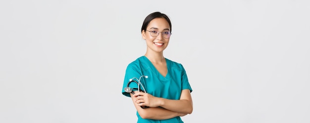 Covid-19, коронавирусная болезнь, концепция медицинских работников. Профессиональный красивый азиатский врач, медицинский работник в очках и скрабах, скрещенные руки и улыбающийся, белый фон.
