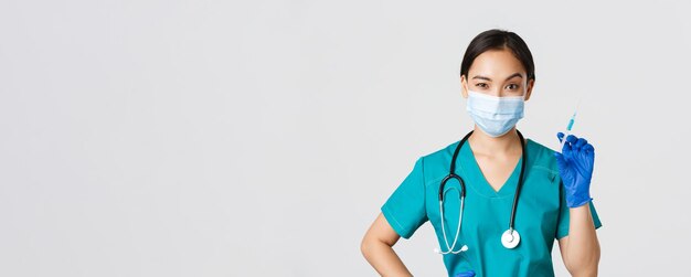 Covid-19, coronavirus disease, healthcare workers concept. Sassy cute asian female nurse, intern in medical mask and gloves holding syringe with vaccine, ready to make shot, white background.