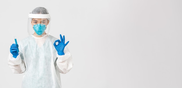 Free photo covid-19, coronavirus disease, healthcare workers concept. serious asian female doctor, professional researcher in personal protective equipment showing okay gesture, holding test-tube with vaccine