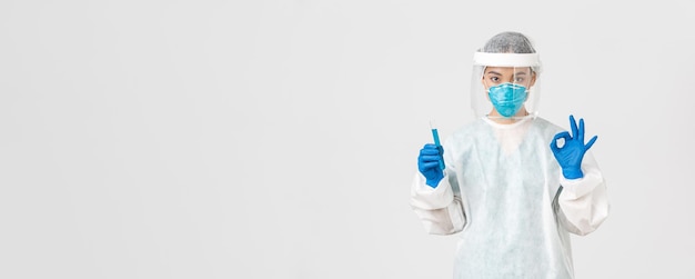 Free photo covid-19, coronavirus disease, healthcare workers concept. serious asian female doctor, professional researcher in personal protective equipment showing okay gesture, holding test-tube with vaccine