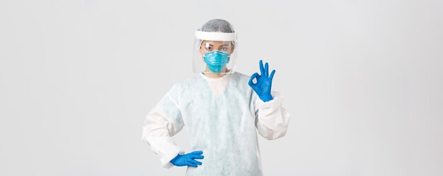 Covid-19, coronavirus disease, healthcare workers concept. Serious-looking professional female asian doctor in personal protective equipment, showing okay gesture, ensure safety of patient.
