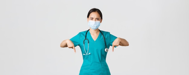 Covid-19, coronavirus disease, healthcare workers concept. Serious-looking worried asian female doctor inform patients, physician in medical mask and scrubs frowning, pointing fingers down