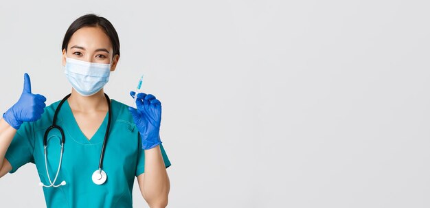 Ковид-19, коронавирусная болезнь, концепция медицинских работников. Улыбающийся красивый азиатский медицинский работник, медсестра в маске и перчатках с большим пальцем вверх, держит шприц с вакциной, стоит на белом фоне