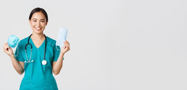 Covid-19, coronavirus disease, healthcare workers concept. Smiling pretty asian female nurse, physician in scrubs showing respirator and medical mask, suggest patients ways of virus protection