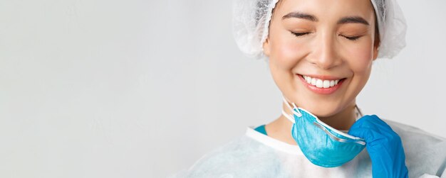 Foto gratuita covid-19, malattia da coronavirus, concetto di operatori sanitari. respiratore da decollo per dottoressa asiatica sorridente sorridente e con danni alla pelle causati dai dispositivi di protezione individuale, sfondo bianco