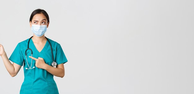 Covid-19, coronavirus disease, healthcare workers concept. Suspicious or doubtful asian female doctor in scrubs and medical mask, looking uncertain and pointing upper left corner, white background