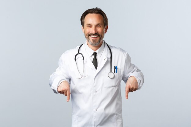 Free photo covid-19, coronavirus outbreak, healthcare workers and pandemic concept. cheerful smiling male doctor in white coat inviting take test in clinic, pointing fingers down, advertising