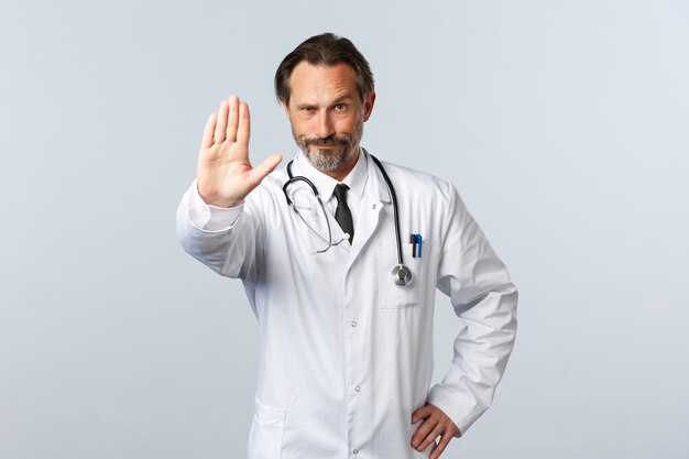 Covid-19, coronavirus outbreak, healthcare workers and pandemic concept. Displeased serious doctor in white coat, extend arm to show stop sign, scolding or giving warning, disapprove action
