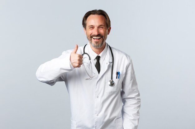 Covid-19, coronavirus outbreak, healthcare workers and pandemic concept. Handsome cheerful male doctor in white coat show thumbs-up in approval, agree or like product, recommending