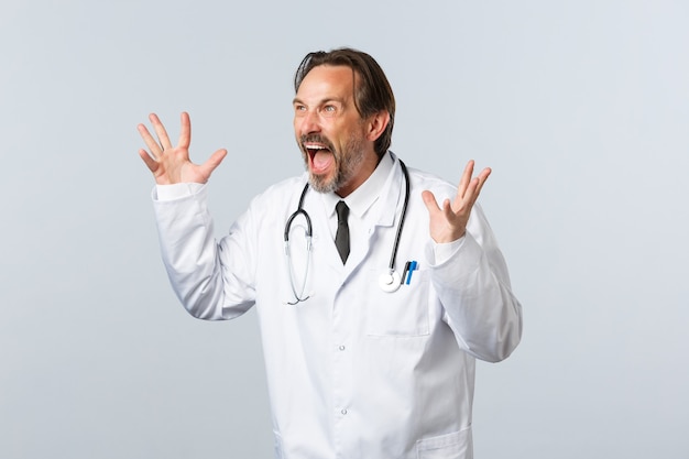 Free photo covid-19, coronavirus outbreak, healthcare workers and pandemic concept. mad crazy doctor in white coat gesturing, confronting employee at clinic, gesturing angry and look left furious