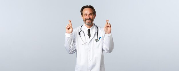 Covid-19, вспышка коронавируса, медицинские работники и концепция пандемии. Оптимистичный улыбающийся врач, верящий в качество услуг клиники, скрестив пальцы на удачу, загадывая желание.