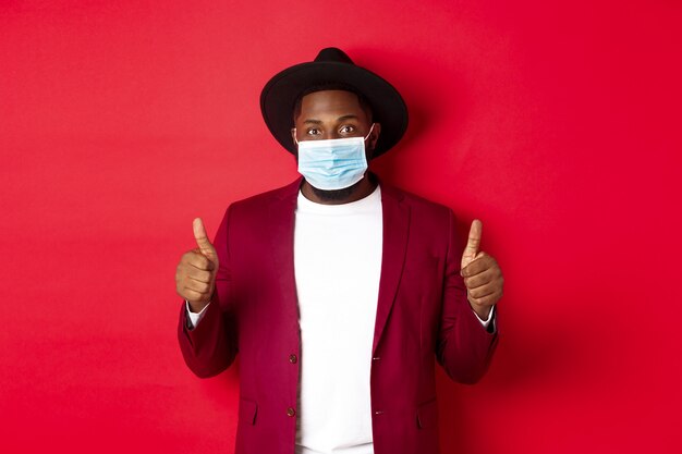 Covid-19 and fashion concept. Stylish african american man in hat and blazer, wearing face mask and showing thumb up, standing over red background.