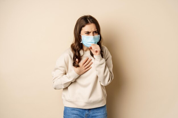 Covid-19 and health concept. Sick woman in medical face mask coughing, feeling ill with sour throat, standing over beige background