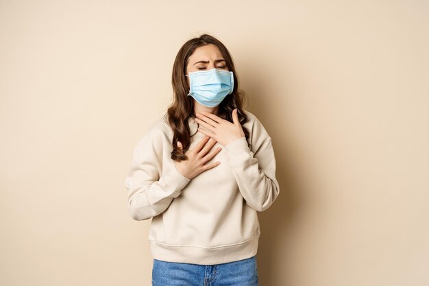 Covid-19 and health concept. Sick woman in medical face mask coughing, feeling ill with sour throat, standing over beige background