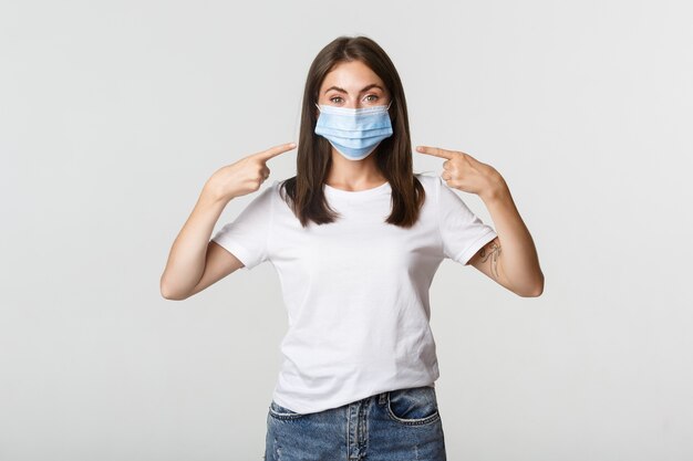 Covid-19, концепция здравоохранения и социального дистанцирования. Привлекательная девушка брюнет в медицинской маске указывая пальцем на лицо, белое.