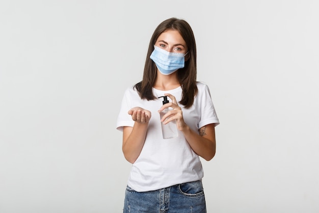 Covid-19, health and social distancing concept. Attractive young brunette woman in medical mask applying hand sanitizer on hand, white.